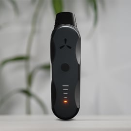 Vaporizador de hierbas AirVape XS Go de pie sobre una superficie blanca con LED naranja encendido y planta desenfocada al fondo.