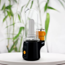 Un elegante Bomb Sykloud E Rig negro con una copa de cristal y una cámara de calentamiento recubierta de naranja, mostrado sobre una superficie blanca con un fondo difuminado de hojas verdes.